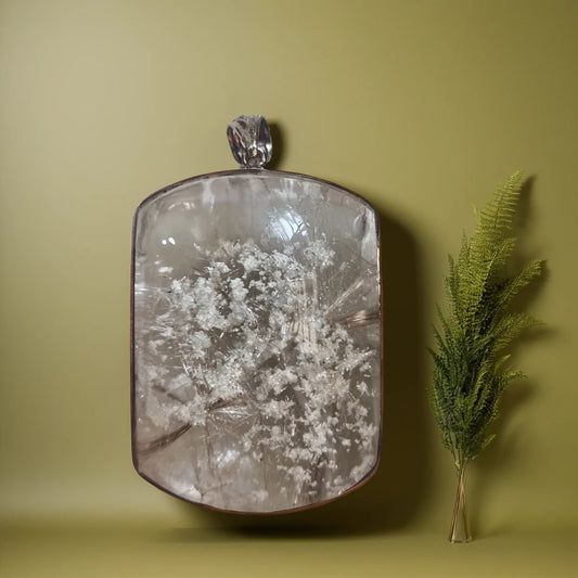 A Garden Quartz with Rutile Silver Pendant displayed against a neutral background, with a green plant to the side.