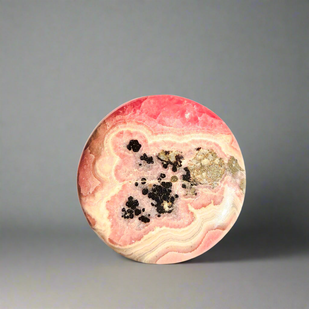 A polished rhodochrosite coin slab with bands of pink and red color, showcasing black and white inclusions.