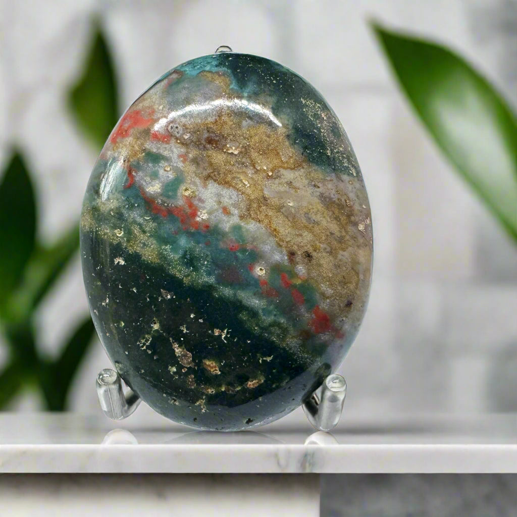 A polished Blood Stone palm stone with green base color and red spots, displayed on a stand.