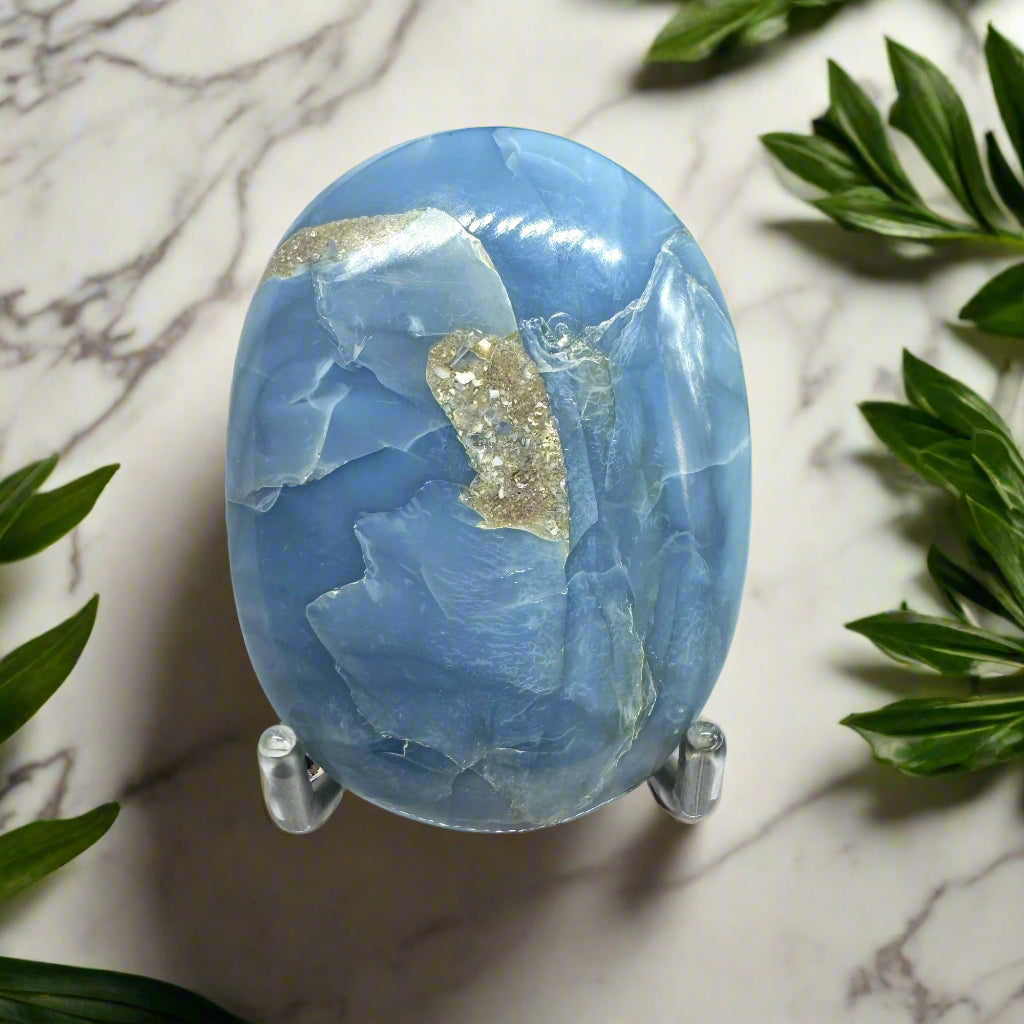 A polished Blue Opal palm stone displayed on a stand, with a marble background and green leaves around it.