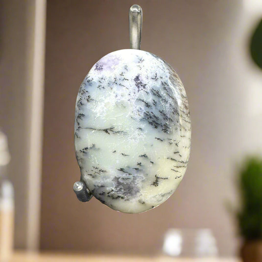 A polished Dendrite Opal palm stone with distinctive fern-like patterns, suspended by a loop, displayed against a blurred background.