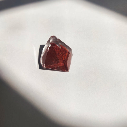A red phantom quartz freeform crystal with triangular shape and polished surface, placed on a light background casting a shadow.