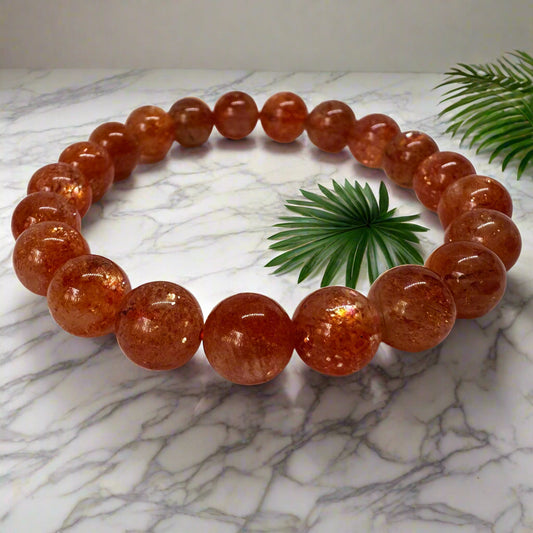 A bracelet made of orange sunstone beads displayed on a marble surface with a green leaf in the background.