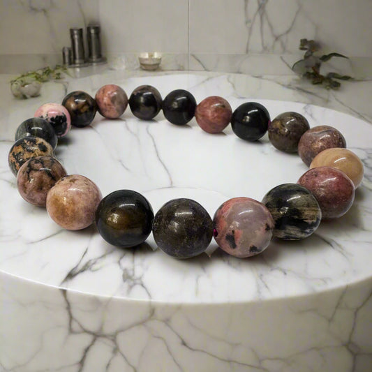 A Rhodonite Bracelet displayed on a marble surface, consisting of round beads with varying shades of pink and black, and some with swirls of black manganese oxide.