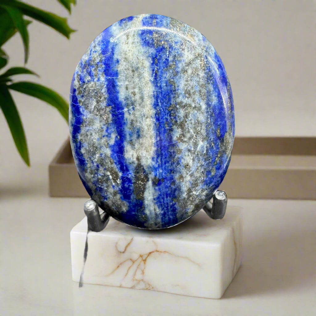 A polished Natural Lapis Lazuli palm stone with deep blue hue and golden flecks, displayed on a white marble stand.