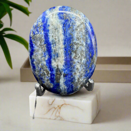 A polished Natural Lapis Lazuli palm stone with deep blue hue and golden flecks, displayed on a white marble stand.