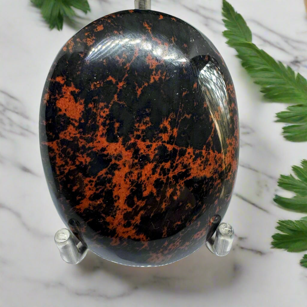 A polished round mahogany palm stone with a rich, earthy red hue, displayed on a stand with a marble background and some green leaves.