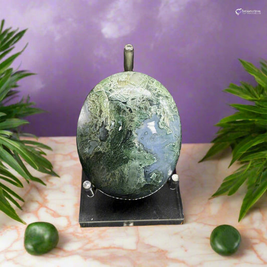 A polished Moss Agate Palmstone displayed on a stand, with two smaller moss agate stones to the left on a marble surface with a purple background and green foliage.