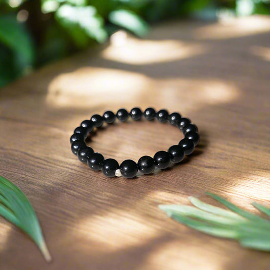 A black tourmaline bracelet with 8mm beads, placed on a wooden surface surrounded by green foliage and a shadow cast over it.