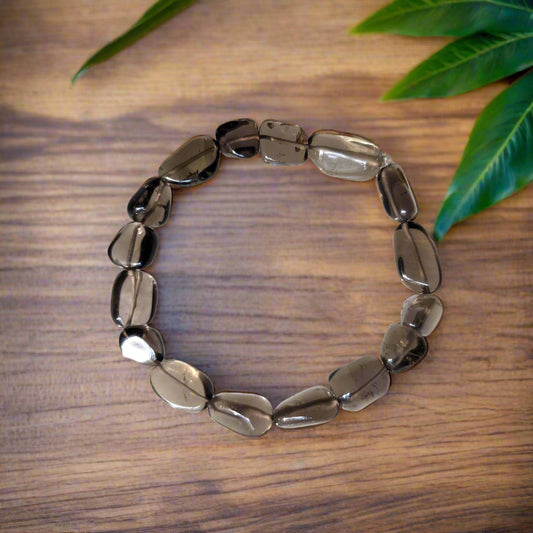 A smokey quartz tumble bracelet displayed on a wooden surface with a few green leaves in the background.