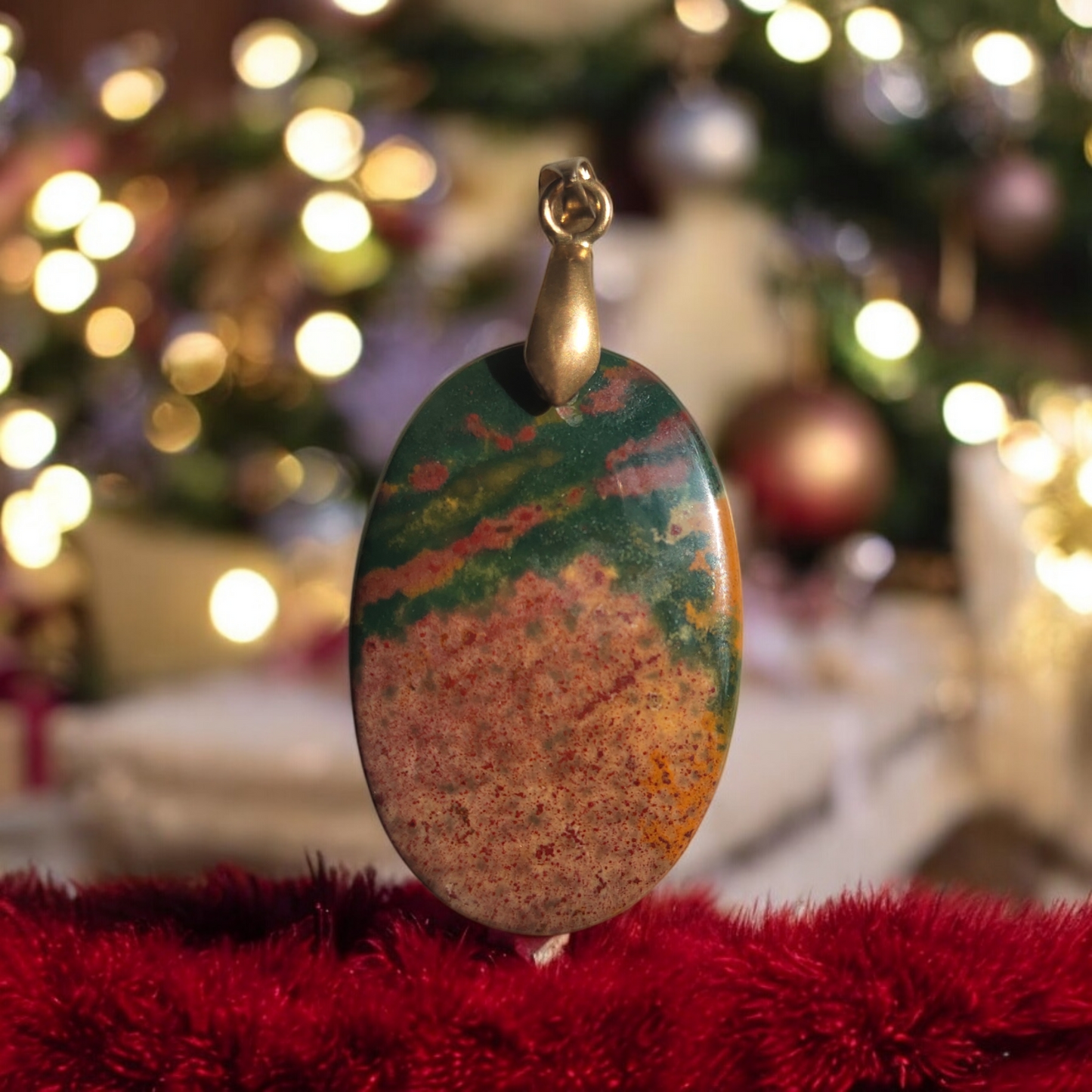 An oval-shaped bloodstone pendant with a mix of green and red colors, displayed in front of a Christmas tree.