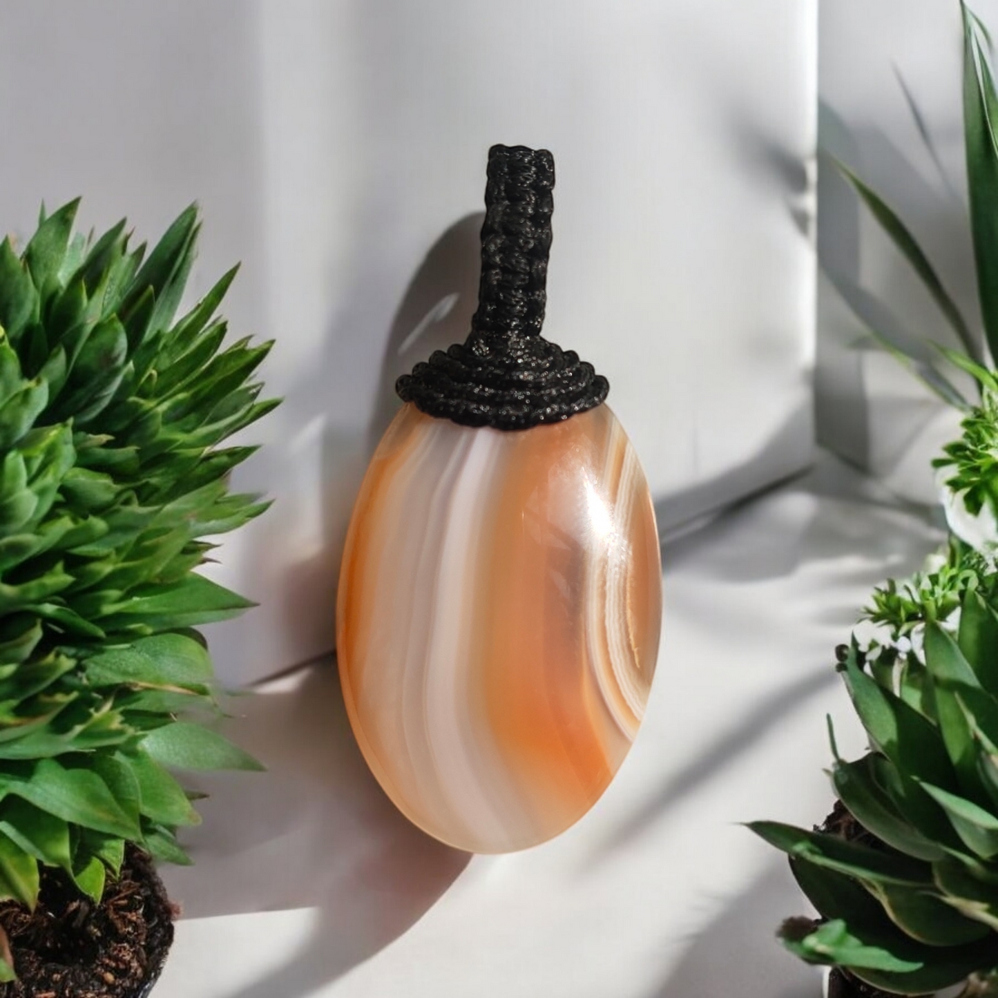 A red carnelian thread pendant displayed against a white background with green plants to the sides.