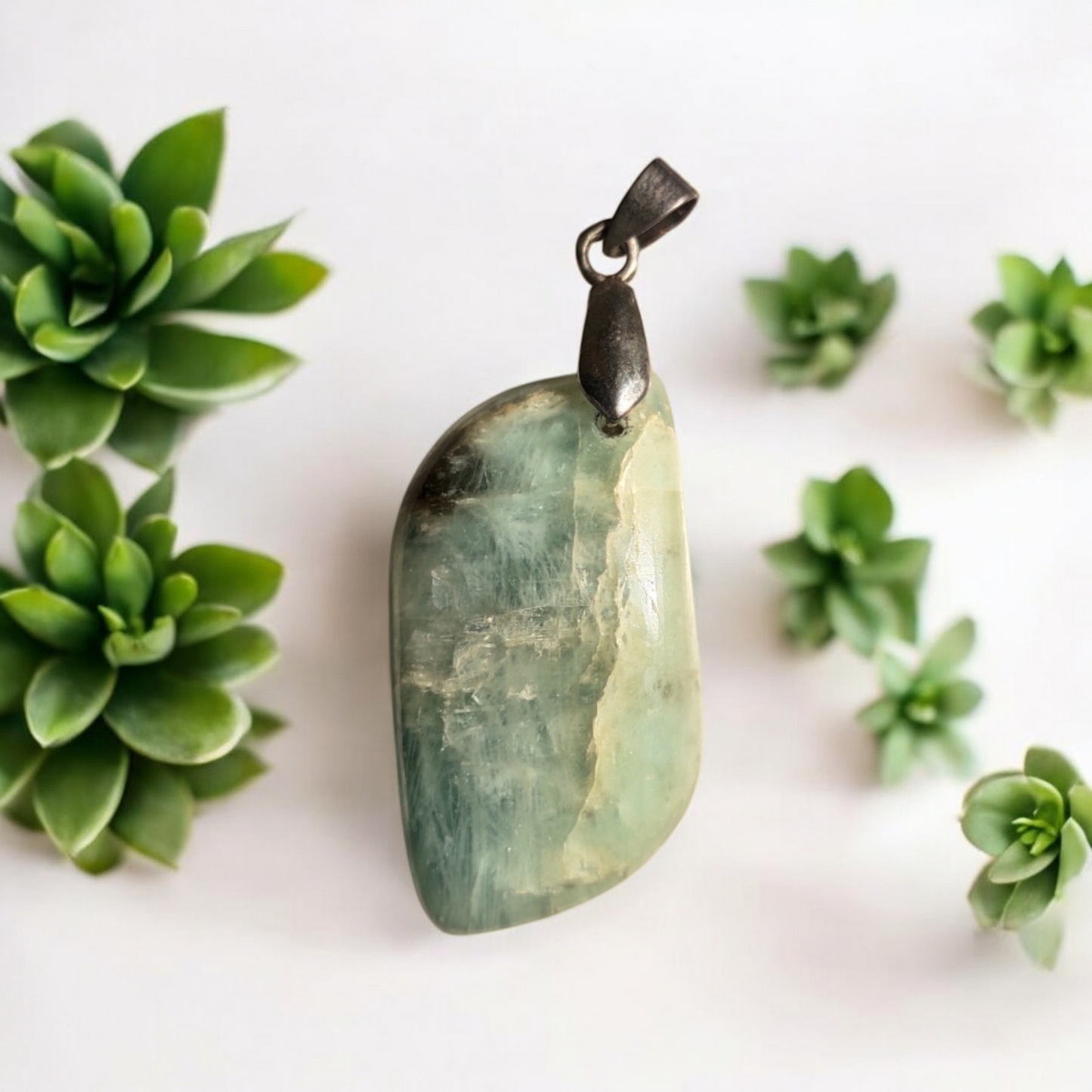 An aquamarine leaf pendant with a silver-colored loop and chain, displayed against a white background with green succulent plants to the side.