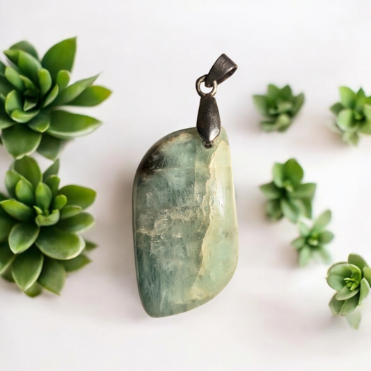 An aquamarine leaf pendant with a silver-colored loop and chain, displayed against a white background with green succulent plants to the side.